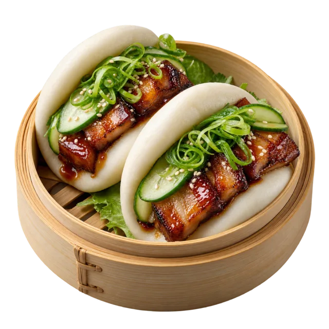 Two white folded bao buns holding glazed meat, cucumber slices and green garnish in a bamboo steamer