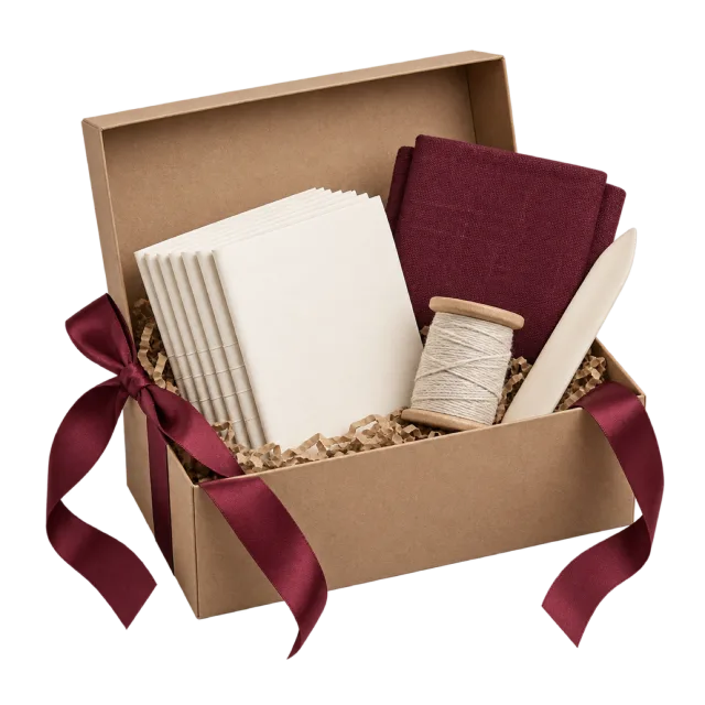 An open kraft box containing folded paper, a burgundy book cover and a spool of thread